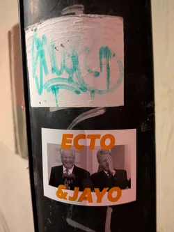 Street sticker The sticker features a white rectangular background with two black and white photographic portraits of political figures. On the left, Boris Yeltsin is depicted smiling broadly, standing behind a microphone. On the right, Bill Clinton is shown with his hand covering his face in a 'facepalm' gesture. Above the images, in large orange capital letters, is the word 'ECTO', and below the images, also in large orange capital letters, is '& JAYO'. The sticker is applied to a dark, textured pole and shows some minor wrinkles and wear.