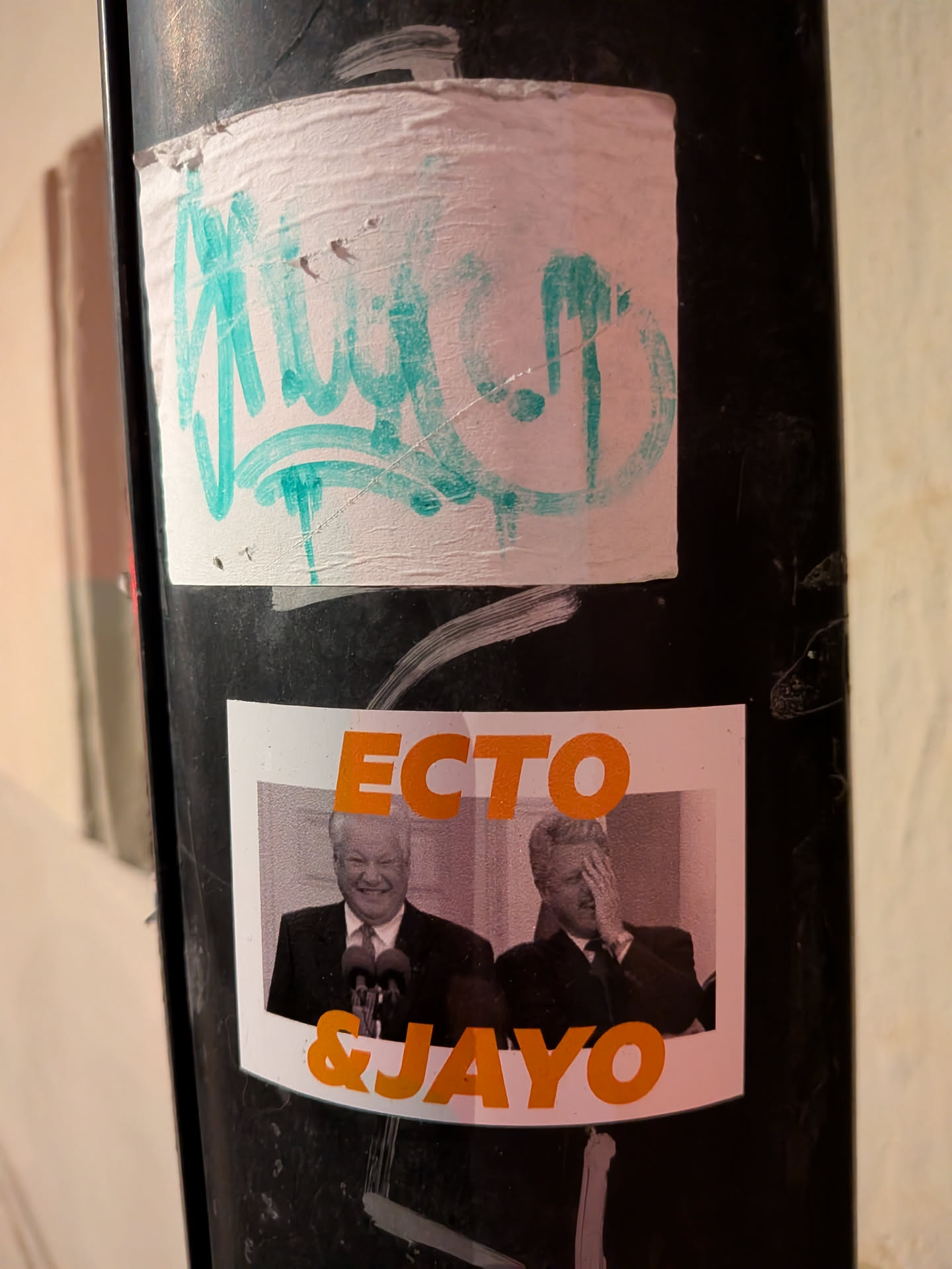 The sticker features a white rectangular background with two black and white photographic portraits of political figures. On the left, Boris Yeltsin is depicted smiling broadly, standing behind a microphone. On the right, Bill Clinton is shown with his hand covering his face in a 'facepalm' gesture. Above the images, in large orange capital letters, is the word 'ECTO', and below the images, also in large orange capital letters, is '& JAYO'. The sticker is applied to a dark, textured pole and shows some minor wrinkles and wear.