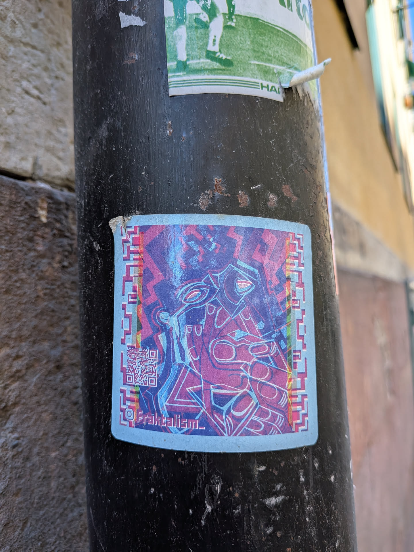 A rectangular sticker with slightly rounded corners, featuring a complex, abstract design dominated by shades of purple, fuchsia, and light blue. The central artwork consists of intertwined, flowing lines and geometric shapes that suggest a stylized, almost alien-like face or creature, with prominent eye-like elements. The background is a darker, textured purple. A distinctive border surrounds the central image, composed of alternating blocks of pink, blue, and purple, creating a pixelated or 'glitch' effect, particularly noticeable on the vertical sides. In the bottom left corner, there is a readable QR code. Below the main artwork, centered at the bottom, is a social media handle. The sticker is affixed to a dark, weathered metal pole.