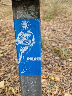 Street sticker A blue sticker affixed to a metal post depicts a white stencil of a person, possibly a soccer player, holding a ball. The style suggests street art or graffiti.  The text 'DU STIM' is partially visible at the bottom. The sticker appears slightly damaged or worn.
