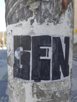 Street sticker A black and white sticker with the word "GEN" written in bold, uppercase letters. The sticker is applied to a weathered, light gray surface, possibly concrete, with visible signs of wear and tear, such as peeling and discoloration. The sticker itself shows some signs of wear but the text remains clearly visible.