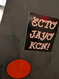 Street sticker The sticker is rectangular with a black background. It features three lines of text in a bold, stylized font. The letters are filled with white and have a prominent red outline, giving them a raised, almost 3D appearance. The text reads 'ECTO', 'JAYO', and 'KCN!' from top to bottom. There is a faint red smudge above the 'ECTO' text.