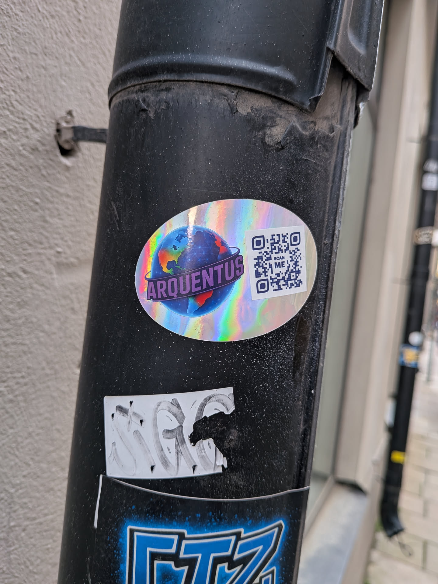 An oval holographic sticker on a dark pole. It depicts a colorful globe encircled by a purple banner with the text 'ARQUENTUS', alongside a QR code labeled 'SCAN ME'.