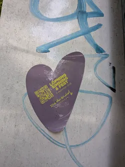 Street sticker A heart-shaped purple sticker on a concrete wall with blue graffiti. Features yellow text for 'LÖPNING SIMNING & FEST', 'Dockside! SWIM & GRAVEL RUN', a date, and a QR code.