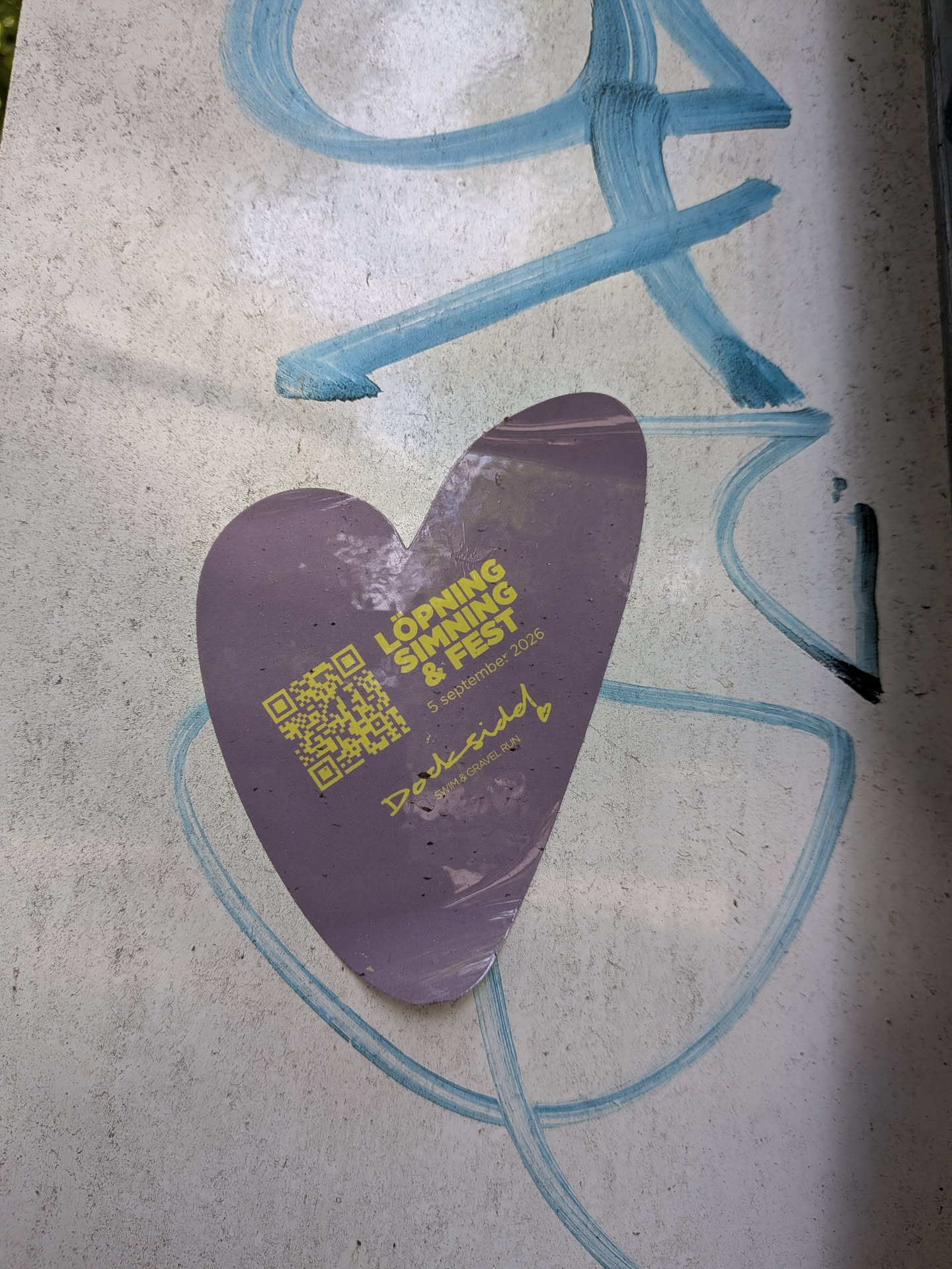 A heart-shaped purple sticker on a concrete wall with blue graffiti. Features yellow text for 'LÖPNING SIMNING & FEST', 'Dockside! SWIM & GRAVEL RUN', a date, and a QR code.