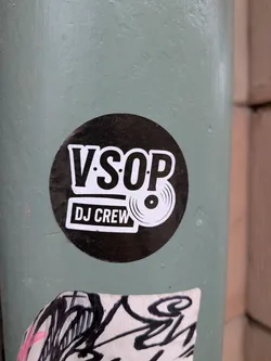 A circular sticker featuring a bold, black background. The main text 'V.SOP' is prominently displayed in large, white, sans-serif capital letters with a subtle outline. Below 'V.SOP', the words 'DJ CREW' are written in smaller, white, sans-serif capital letters, enclosed within a white rectangular box with rounded corners. To the right of 'DJ CREW' and slightly overlapping the 'P' in 'V.SOP', there is a white graphic icon of a vinyl record. The overall design is clean, minimalist, and uses a high-contrast black and white color scheme.