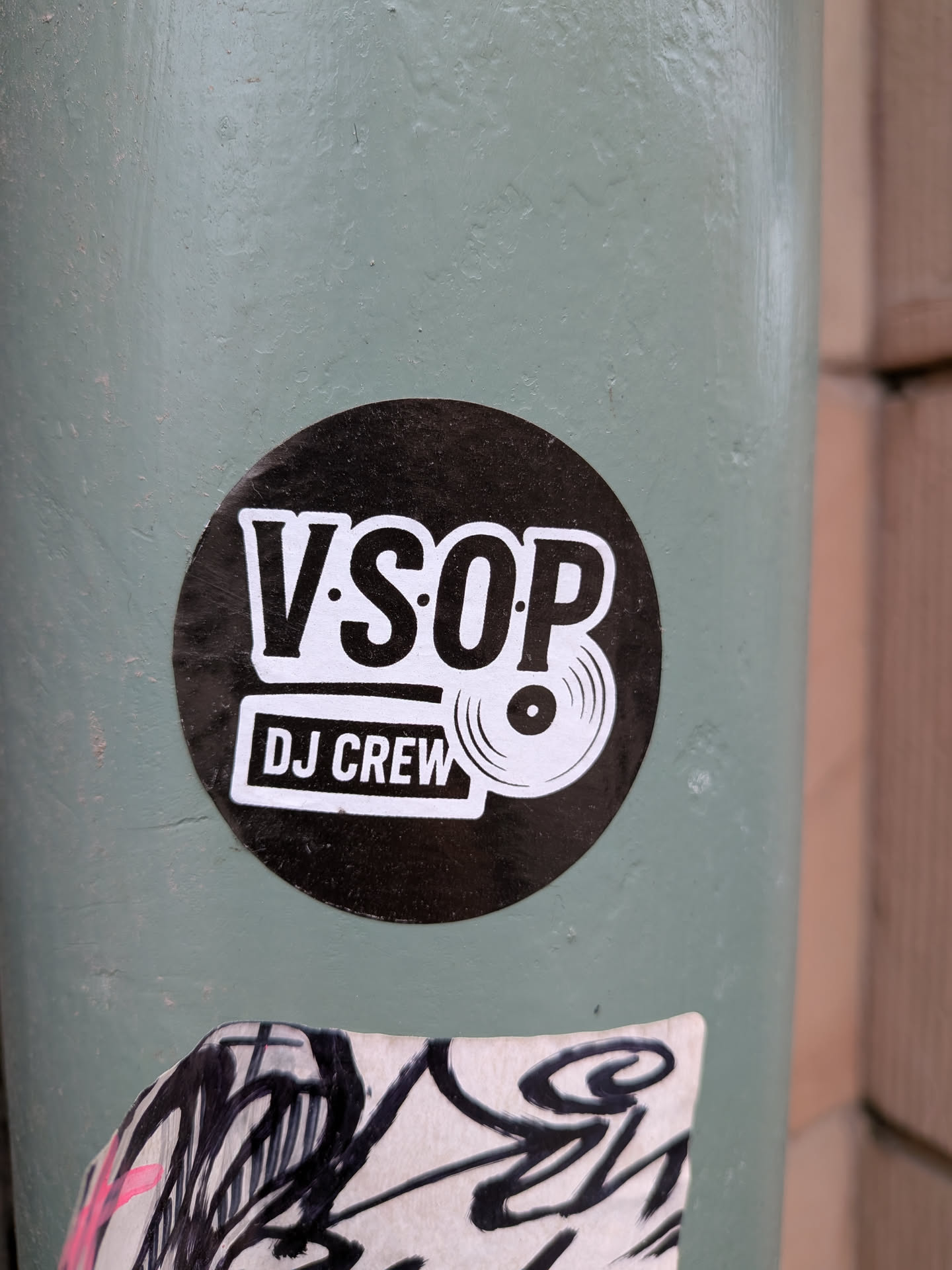 A circular sticker featuring a bold, black background. The main text 'V.SOP' is prominently displayed in large, white, sans-serif capital letters with a subtle outline. Below 'V.SOP', the words 'DJ CREW' are written in smaller, white, sans-serif capital letters, enclosed within a white rectangular box with rounded corners. To the right of 'DJ CREW' and slightly overlapping the 'P' in 'V.SOP', there is a white graphic icon of a vinyl record. The overall design is clean, minimalist, and uses a high-contrast black and white color scheme.