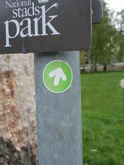 Street sticker A small, round, green sticker with a white upward-pointing arrow is affixed to a gray metal pole. The sticker appears to be a directional marker.  A sign above it reads 'National Stadspark'.