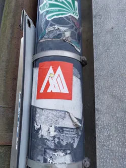 A square sticker with a red background and a stylized white geometric mountain logo.  The sticker appears to be affixed to a pole with other stickers and markings.