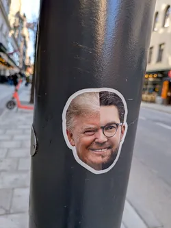 The sticker is an oval-shaped, photographic composite portrait with a clean white border, affixed to a dark grey pole. It features the faces of two men seamlessly merged down the vertical center. The left half of the face belongs to an older man with fair, possibly blonde or grey, hair, a ruddy complexion, and a prominent chin. He is depicted winking with his left eye and smiling broadly, revealing teeth. The right half of the face belongs to a younger man with dark, neatly combed hair, a well-groomed beard, and dark-rimmed round glasses. He is smiling with his right eye open, also revealing teeth. The two halves are expertly blended to create a single, cohesive, smiling and winking expression.