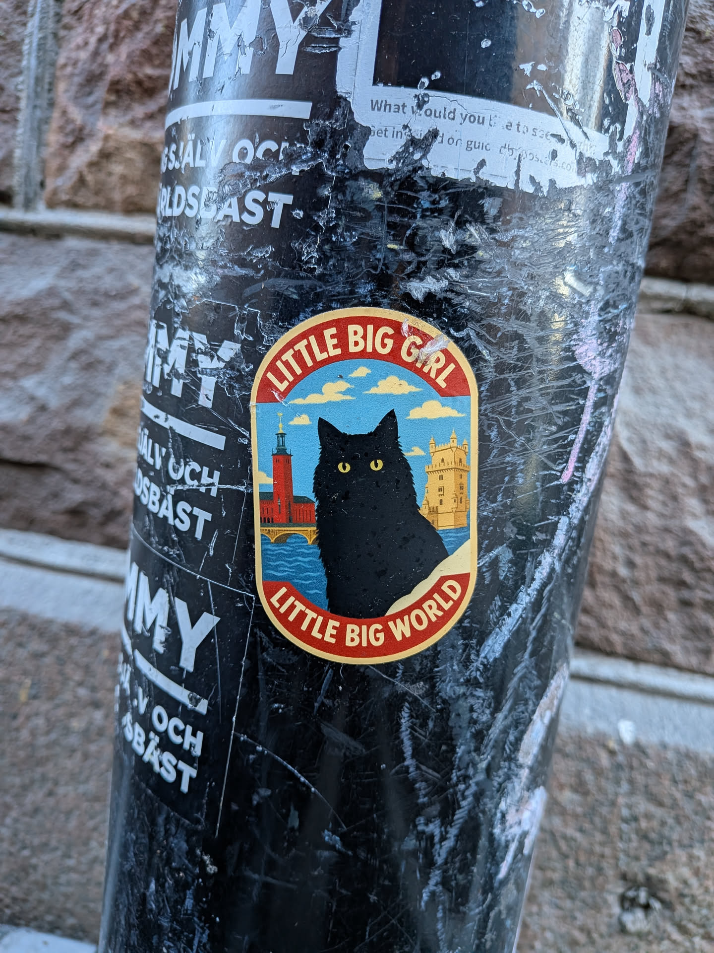 The sticker is oval-shaped with a prominent red outer border and a thinner yellow inner border. The central image features a large, fluffy black cat with bright yellow eyes, positioned in the foreground, looking slightly to the right. In the background, on the left, is the red brick tower of Stockholm City Hall, with a bridge extending over blue water. On the right, partially obscured by the cat, is the yellow-stone Belém Tower. The sky above is a light blue with several white, fluffy clouds. The water is depicted with wavy lines in varying shades of blue. The overall style is a cute, illustrative cartoon.