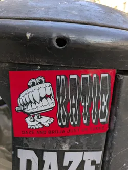 Street sticker The sticker has a red background. On the left is a cartoon drawing of a creature with very large, sharp teeth and small feet. To the right of the drawing is the name "KATIEE" printed in a stylized white font with a black outline. Underneath the drawing is the text "DAZE AND BRUJA JUST US THREE".