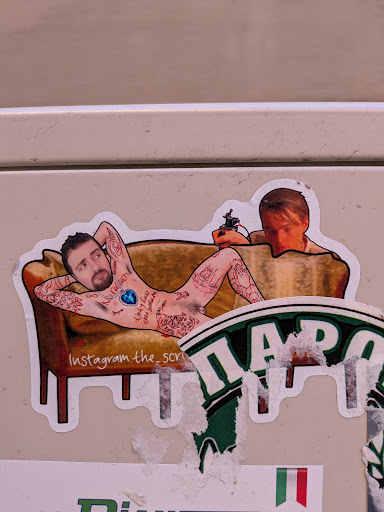 A sticker showing a naked tattooed man lying on a couch being tattooed. It seems to be a collage of an individual's face put on the tattooed model. The sticker includes social media links.