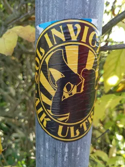 Street sticker This circular sticker boasts a bold design.  The words "Invicta Ulster" are prominently displayed around a central image of a person holding a megaphone, set against a yellow and black striped background.  The sticker is affixed to a gray pole outdoors.