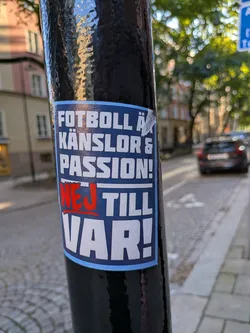 Street sticker The sticker is predominantly blue with white text and a thin light blue border. The text is arranged in multiple lines. The top line reads 'FOTBOLL Ä', followed by 'KÄNSLOR &' on the second line. The third line states 'PASSION!'. Below this, the word 'NEJ' is prominently displayed in red with a red underline, followed by 'TILL' and 'VAR!' in large white capital letters on a blue background. The sticker appears slightly worn and peeling at the edges, suggesting it's been affixed for some time. The overall design is bold and aims to convey a strong message.