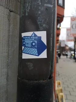 Street sticker A square white sticker featuring a bold blue graphic design. The central image depicts a stylized boombox resting on top of a keyboard, forming a pyramid-like structure. The boombox has distinct speakers, buttons, and a cassette deck area. The keyboard below it shows individual keys. Behind this structure, a large blue triangle points upwards. Above the boombox, the text 'FLORAS KULLE' is written in blue, and below it, 'L.. MASSIVE _ _' is also in blue. The overall aesthetic is minimalist and graphic, using only blue and white colors.