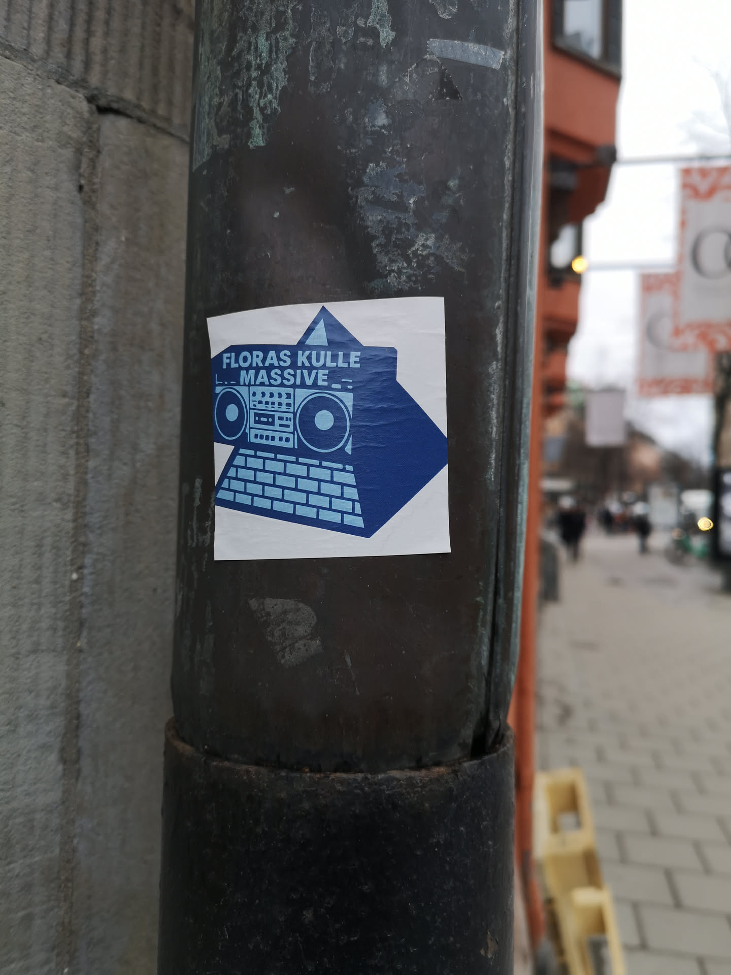 A square white sticker featuring a bold blue graphic design. The central image depicts a stylized boombox resting on top of a keyboard, forming a pyramid-like structure. The boombox has distinct speakers, buttons, and a cassette deck area. The keyboard below it shows individual keys. Behind this structure, a large blue triangle points upwards. Above the boombox, the text 'FLORAS KULLE' is written in blue, and below it, 'L.. MASSIVE _ _' is also in blue. The overall aesthetic is minimalist and graphic, using only blue and white colors.