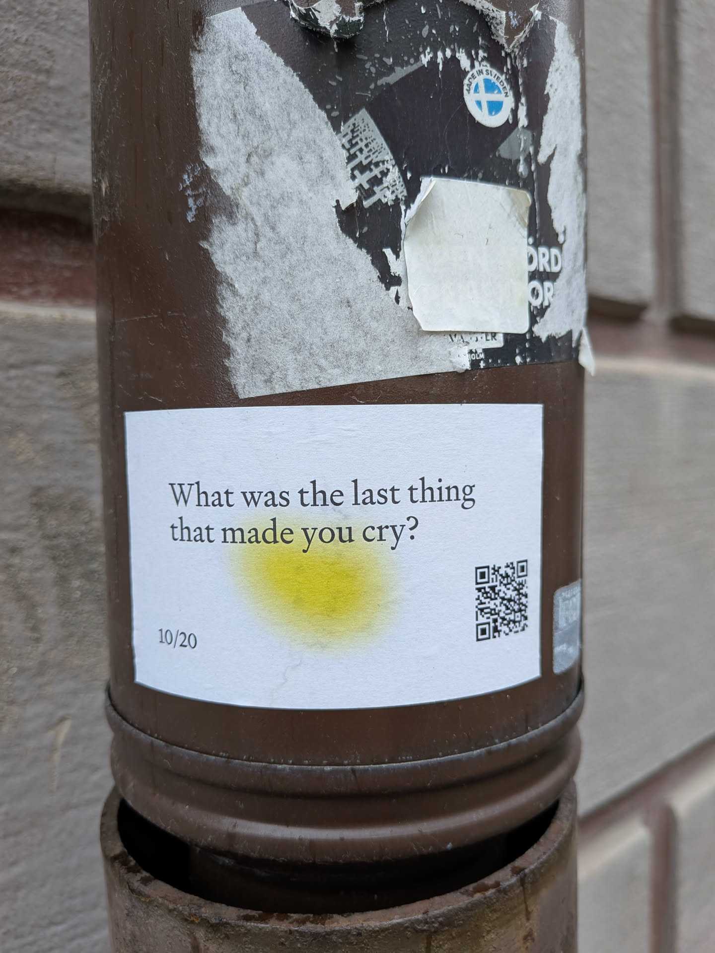 The sticker is a rectangular white label, affixed to a dark brown metal pipe. It features black serif text asking, "What was the last thing that made you cry?". Below this question, to the left, is the number "10/20". A prominent yellow, somewhat translucent, circular smudge is visible in the center of the sticker, partially overlapping the text. To the right, a clear black and white QR code is printed. The edges of the sticker appear slightly worn and lifted in places, indicating it's a physical sticker exposed to elements.
