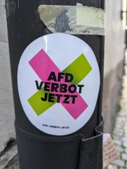 Street sticker A sticker with a pink and green X over the text "AfD Verbot Jetzt", which translates to  "AfD ban now". The bottom of the sticker has a smaller text that reads "afd-verbot.jetzt"
