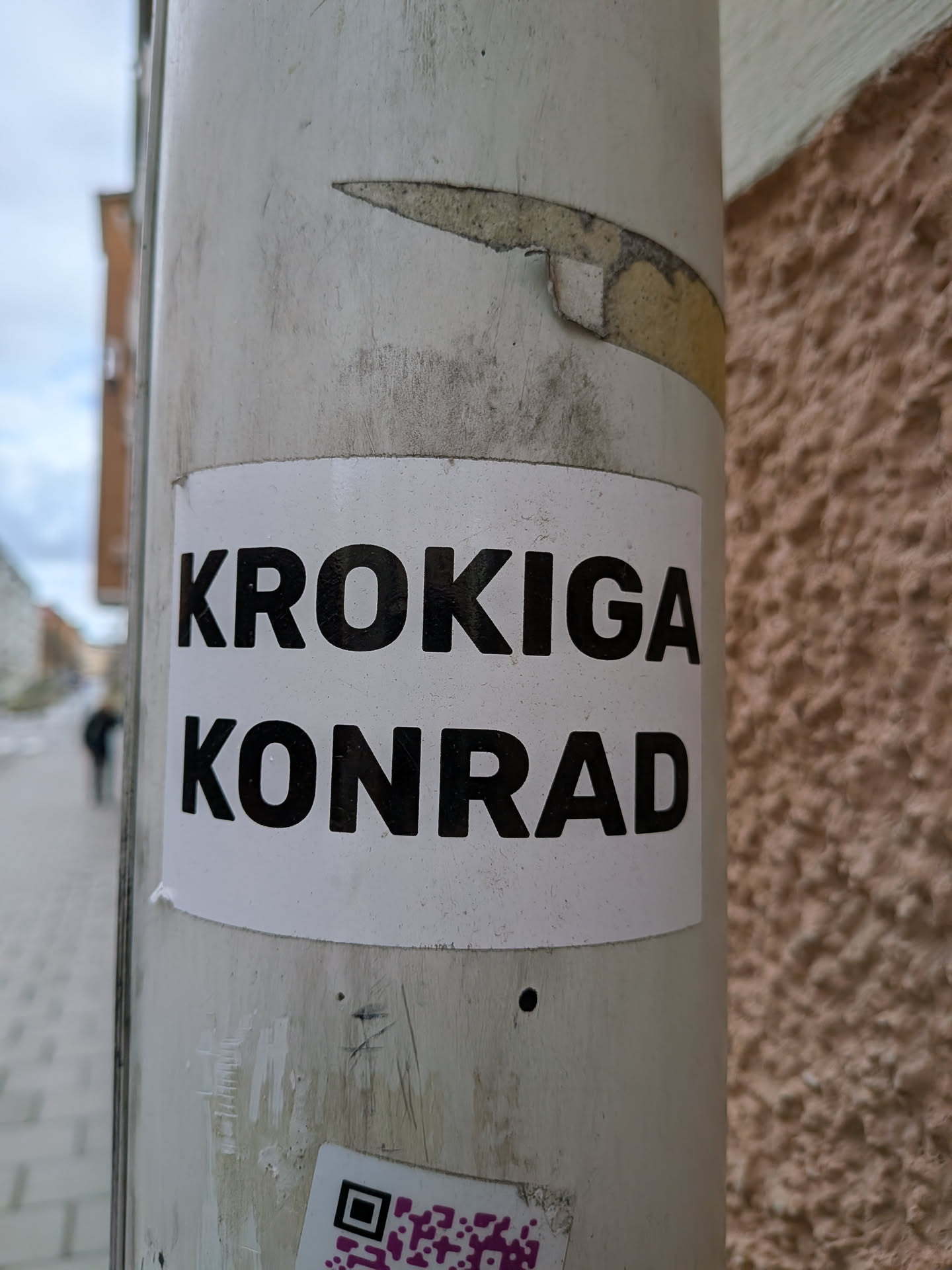 The sticker is rectangular with slightly rounded corners, featuring a clean white background. It displays two lines of text in bold, black, sans-serif uppercase letters. The top line reads 'KROKIGA' and the bottom line reads 'KONRAD'. The sticker appears to be made of paper or vinyl and shows minor signs of wear and dirt, consistent with outdoor placement on a light-colored utility pole. There is a partially visible purple and white QR code sticker below it.