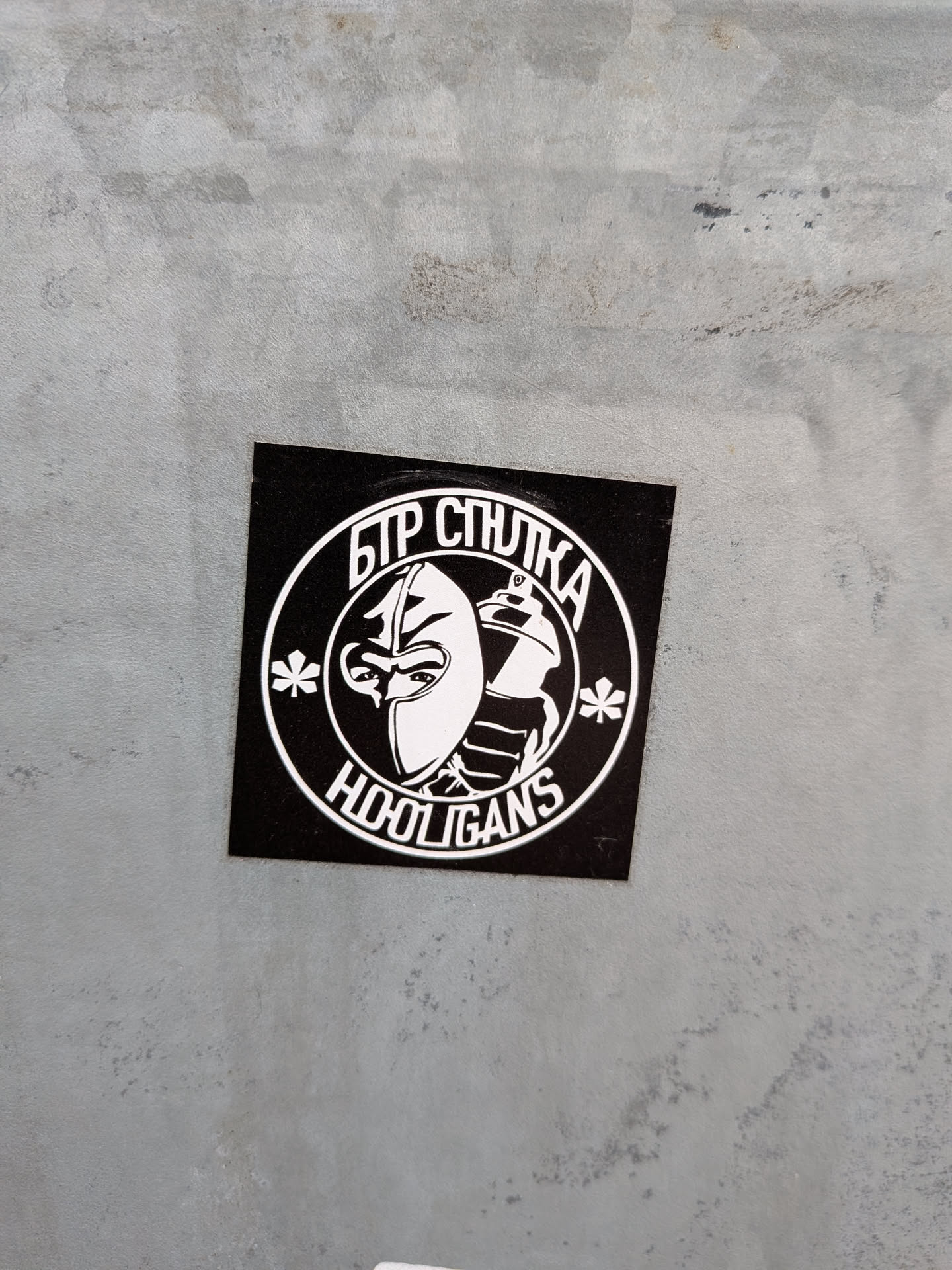 Street sticker The sticker is square with a black background, featuring a prominent white circular emblem in its center. Inside the circle, a stylized design depicts the profile of a face wearing a balaclava or mask on the left, with visible eyes, juxtaposed with a spray paint can on the right. Above this central imagery, within the circle, is Cyrillic text that reads 'БТР СТРЕЛКА'. Below the central design, also within the circle, is the English word 'HOOLIGANS'. Two small asterisk-like symbols flank the central design on either side. The overall aesthetic is stark, high-contrast black and white, giving it a strong, graphic, and somewhat defiant appearance. The sticker is applied to a light grey, textured surface.