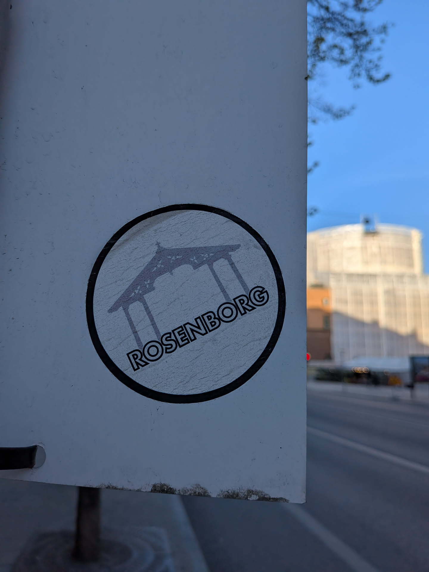 A circular sticker featuring a dark grey silhouette of an architectural structure, likely a pavilion or a section of Rosenborg Castle, against a light grey, textured background. The word 'ROSENBORG' is written in dark grey, uppercase, sans-serif letters along the bottom curve of the circle. The sticker has a distinct black outer border and shows signs of wear and slight creasing.