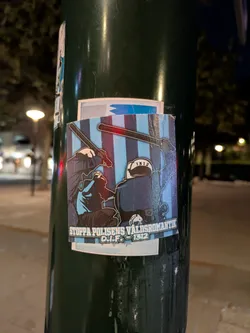 The sticker is rectangular with a slightly irregular white border, affixed to a dark green pole. Its central image depicts two stylized figures in dark blue or black riot gear/uniforms against a background of alternating light blue and darker blue/purple vertical stripes. The figure on the left is seen from behind, holding a dark baton with a distinct red glow emanating from its tip. The figure on the right is facing away, wearing a helmet with '1312' clearly written on it, and holding a large, dark shield. The overall color palette is dominated by blues, black, and red, with white outlines defining the figures. Below the main illustration, there is a white banner containing black text.