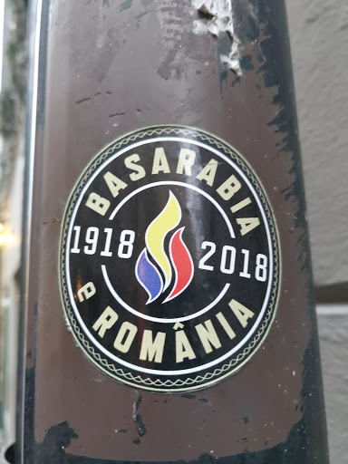 Street sticker A round sticker commemorating the union of Basarabia with Romania in 1918. The sticker features the years 1918 and 2018, a stylized flame in the colors of the Romanian flag, and the text "Basarabia" and "România" around the flame. The sticker is affixed to a brown pole.