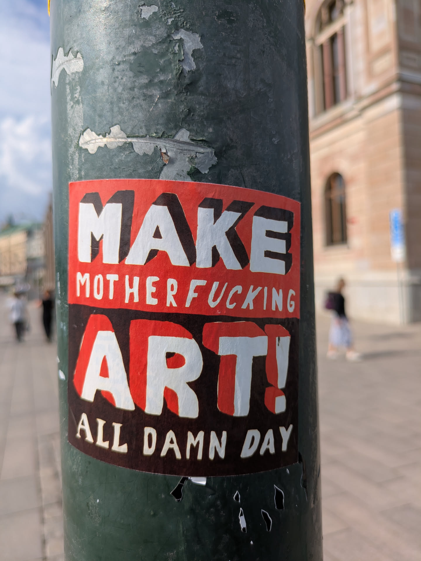 Street sticker A rectangular sticker prominently displayed on a weathered, dark green pole. The sticker features a bold design with a red background for the main text and a dark brown/black background for the secondary text. The words "MAKE" and "ART!" are large, white, block letters with a dark shadow effect, giving them a three-dimensional appearance. Between these two words, in smaller white letters, is "MOTHERFUCKING". Below "ART!", the phrase "ALL DAMN DAY" is written in smaller white sans-serif letters against a dark brown/black strip. The sticker shows signs of wear and slight peeling at the edges.