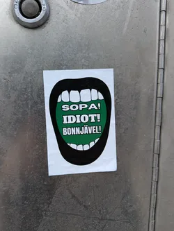 The sticker features a stylized, wide-open mouth with thick black lips and white, blocky teeth. The interior of the mouth is a vibrant, solid green, serving as the background for three lines of bold, white, sans-serif text. The sticker is rectangular with slightly rounded corners, applied to a textured, metallic surface.