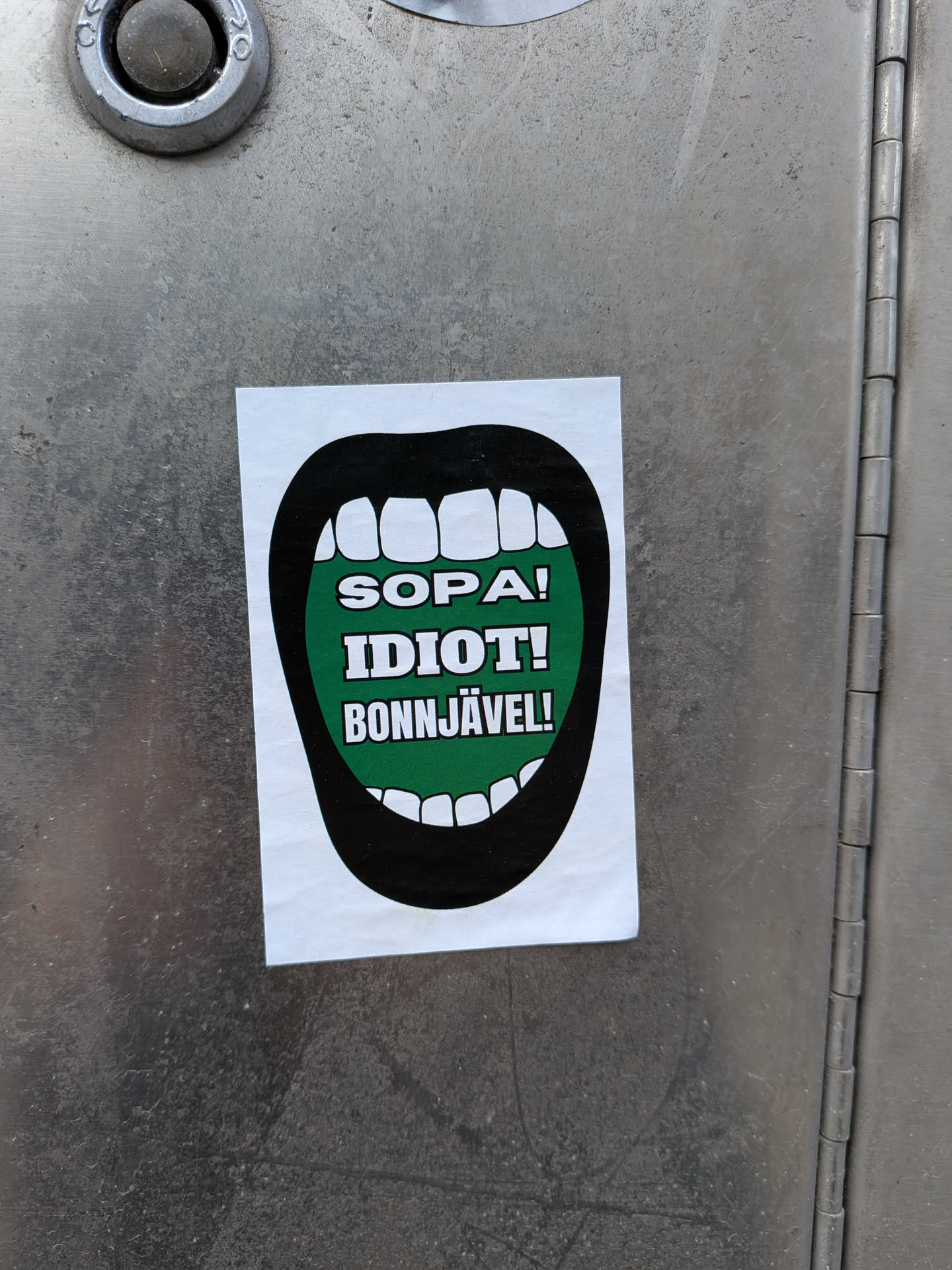 The sticker features a stylized, wide-open mouth with thick black lips and white, blocky teeth. The interior of the mouth is a vibrant, solid green, serving as the background for three lines of bold, white, sans-serif text. The sticker is rectangular with slightly rounded corners, applied to a textured, metallic surface.