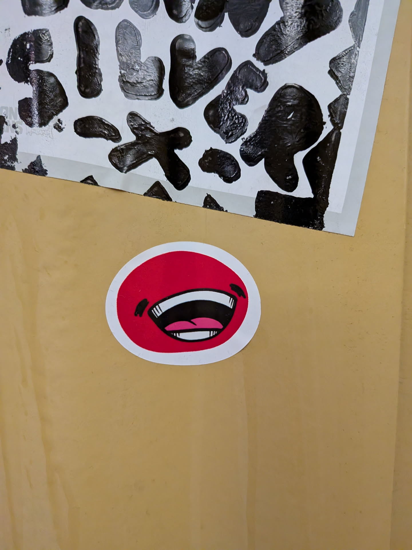 Street sticker The sticker is circular with a thick white border. The inner part is a bright red, depicting an open mouth with a wide, exaggerated smile. The mouth is black with white teeth and a pink tongue. There are also two small black marks above the mouth, resembling eyebrows or cheek marks, adding to its cartoonish and expressive appearance.