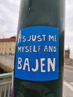 Street sticker A blue square sticker on a green pole, with the text "It's just me myself and Bajen" in white, bold, sans-serif font. The sticker shows slight signs of wear.