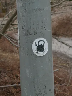 A circular sticker with a white background adhered to a weathered grey metal pole. The central image is a black, somewhat textured silhouette of a kettlebell. To the top-left of the kettlebell, there's a small black heart icon. Curved around the bottom edge of the kettlebell, in black lowercase letters, is the text 'torpetiorhem.se'. The sticker has a slightly worn appearance, particularly on its left edge.
