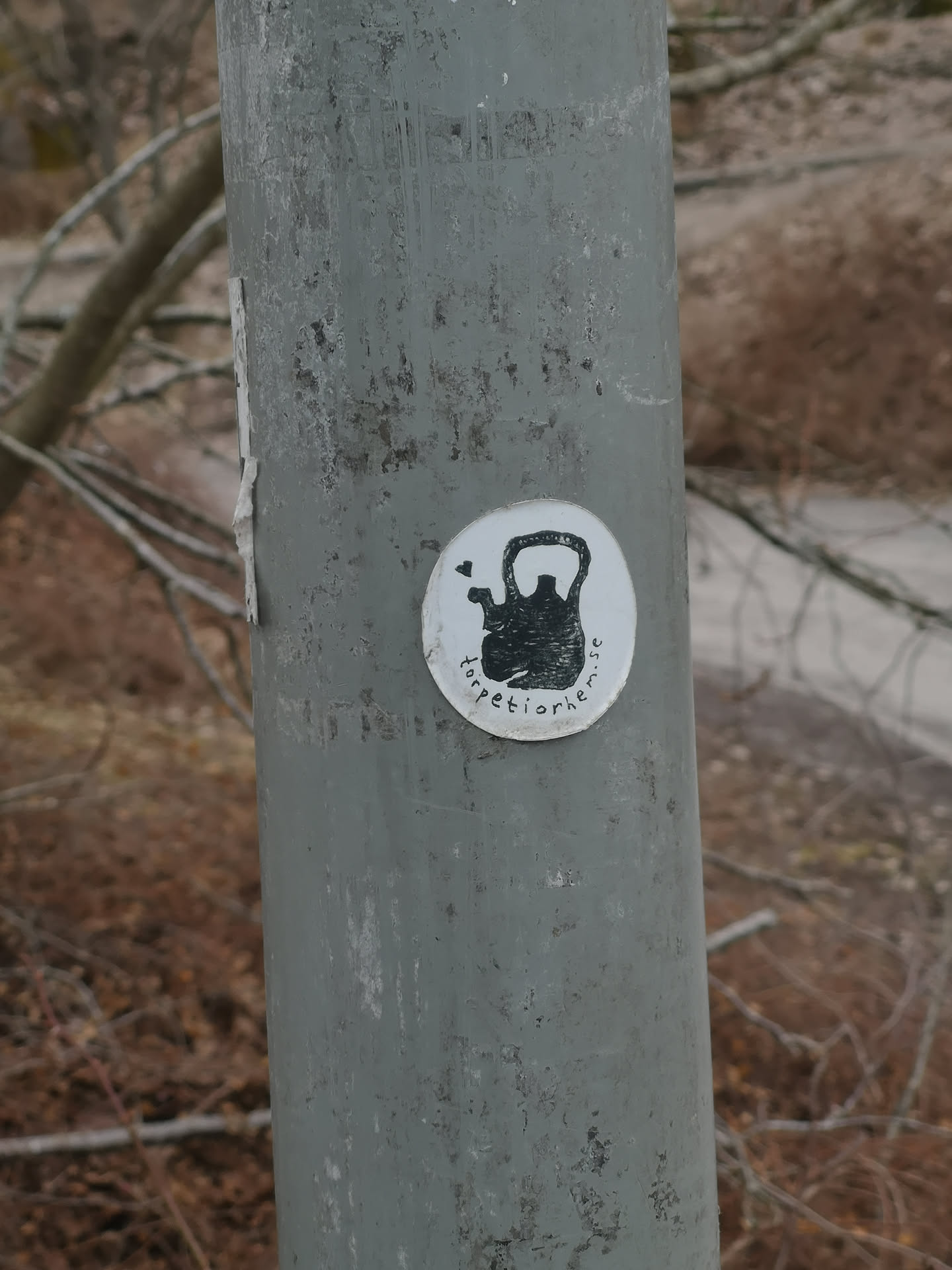 A circular sticker with a white background adhered to a weathered grey metal pole. The central image is a black, somewhat textured silhouette of a kettlebell. To the top-left of the kettlebell, there's a small black heart icon. Curved around the bottom edge of the kettlebell, in black lowercase letters, is the text 'torpetiorhem.se'. The sticker has a slightly worn appearance, particularly on its left edge.