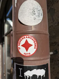 Street sticker A round sticker with a red star and the text "IF Söderkamraterna 1974".  It appears to be related to a Swedish sports club founded in 1974. The sticker is affixed to a brown pole.