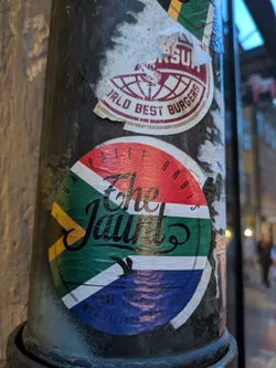 Street sticker The sticker is circular and predominantly features the colors and design of the South African flag. The top segment is red, followed by white, then green, and the bottom is blue. Across the green and white sections, in a stylized black script, is the text "The Jaunt". Above this text, following the curve of the red segment, are the words "PATET ORBIS" in small black capital letters. Below "The Jaunt", in smaller black text, are the words "EST. 2011" and "WWW.THEJAUNT.NET". A silhouette of a bird in flight is also present within the green section.
