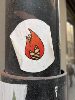 Street sticker This round sticker features a unique design of a corn cob shaped like a flame. The flame is primarily orange and red with black outlines, and the corn kernels are detailed within the flame shape.  It's adhered to a dark-colored pole.