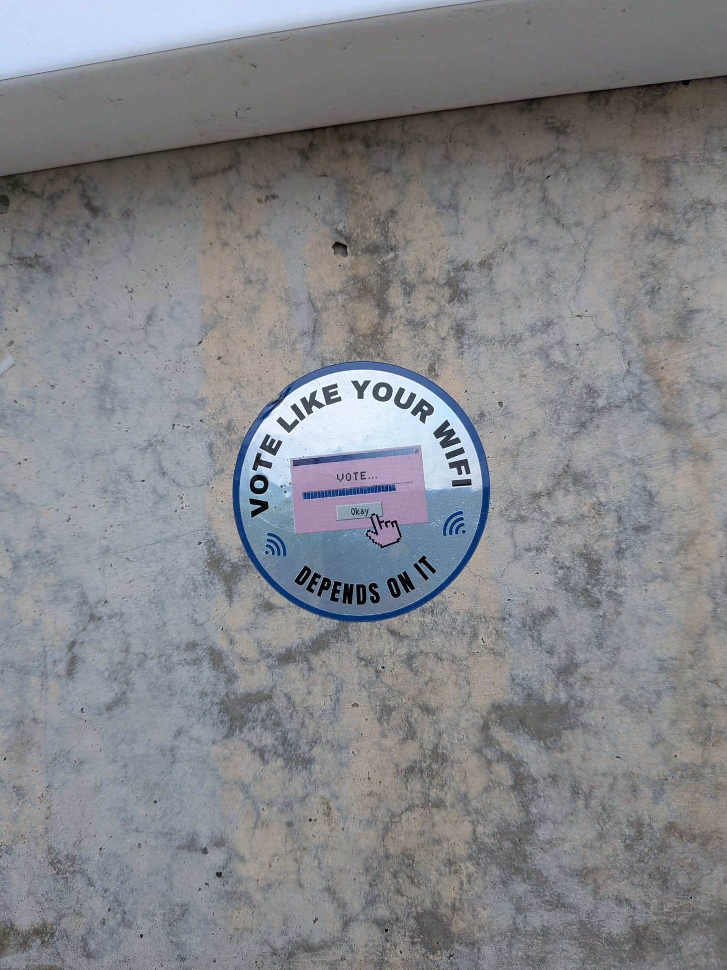 A circular sticker with a blue border adhered to a concrete wall. The sticker features the text 'VOTE LIKE YOUR WIFI' in a bold, black, sans-serif font arched along the top, and 'DEPENDS ON IT' along the bottom. In the center, there is a retro Windows-style dialog box in pink with a loading bar and the text 'VOTE...'. A pixelated hand cursor is clicking an 'Okay' button. Two blue Wi-Fi signal icons are positioned on the left and right sides of the central dialog box.