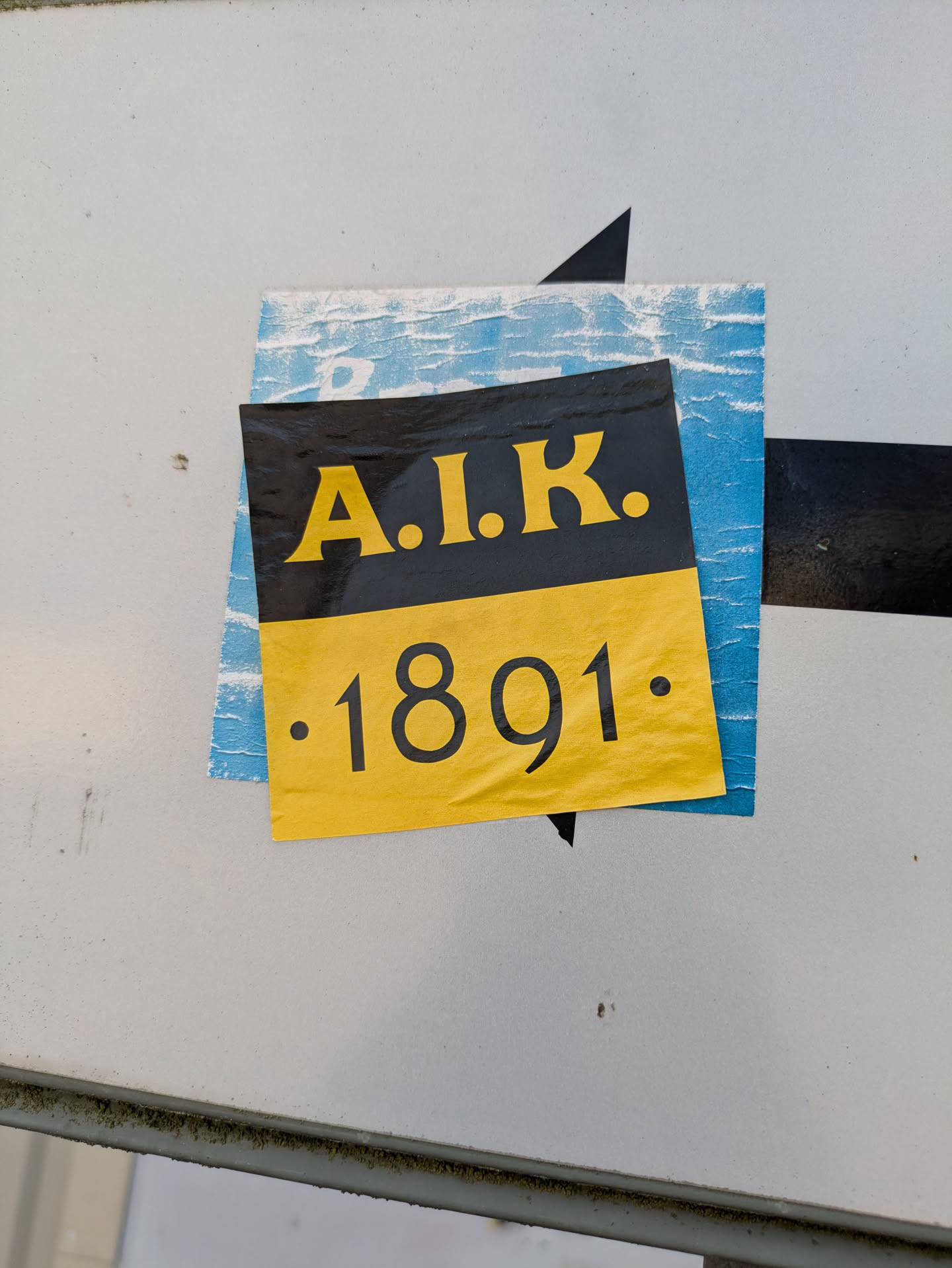 The image shows a square sticker on a white surface. The sticker is divided into two parts: the upper part is black and displays 'A.I.K.' in yellow, while the lower part is yellow and shows '.1891.' in black. Another sticker is partially visible behind it. The main sticker is slightly wrinkled and peeling off.