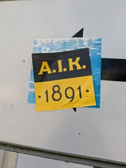 Street sticker The image shows a square sticker on a white surface. The sticker is divided into two parts: the upper part is black and displays 'A.I.K.' in yellow, while the lower part is yellow and shows '.1891.' in black. Another sticker is partially visible behind it. The main sticker is slightly wrinkled and peeling off.