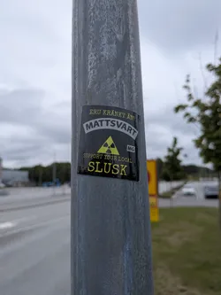 Street sticker Black rectangular sticker on a metal pole. Features yellow and white text in Swedish and English, centered around a yellow radioactive symbol with 'MG' in a small box.