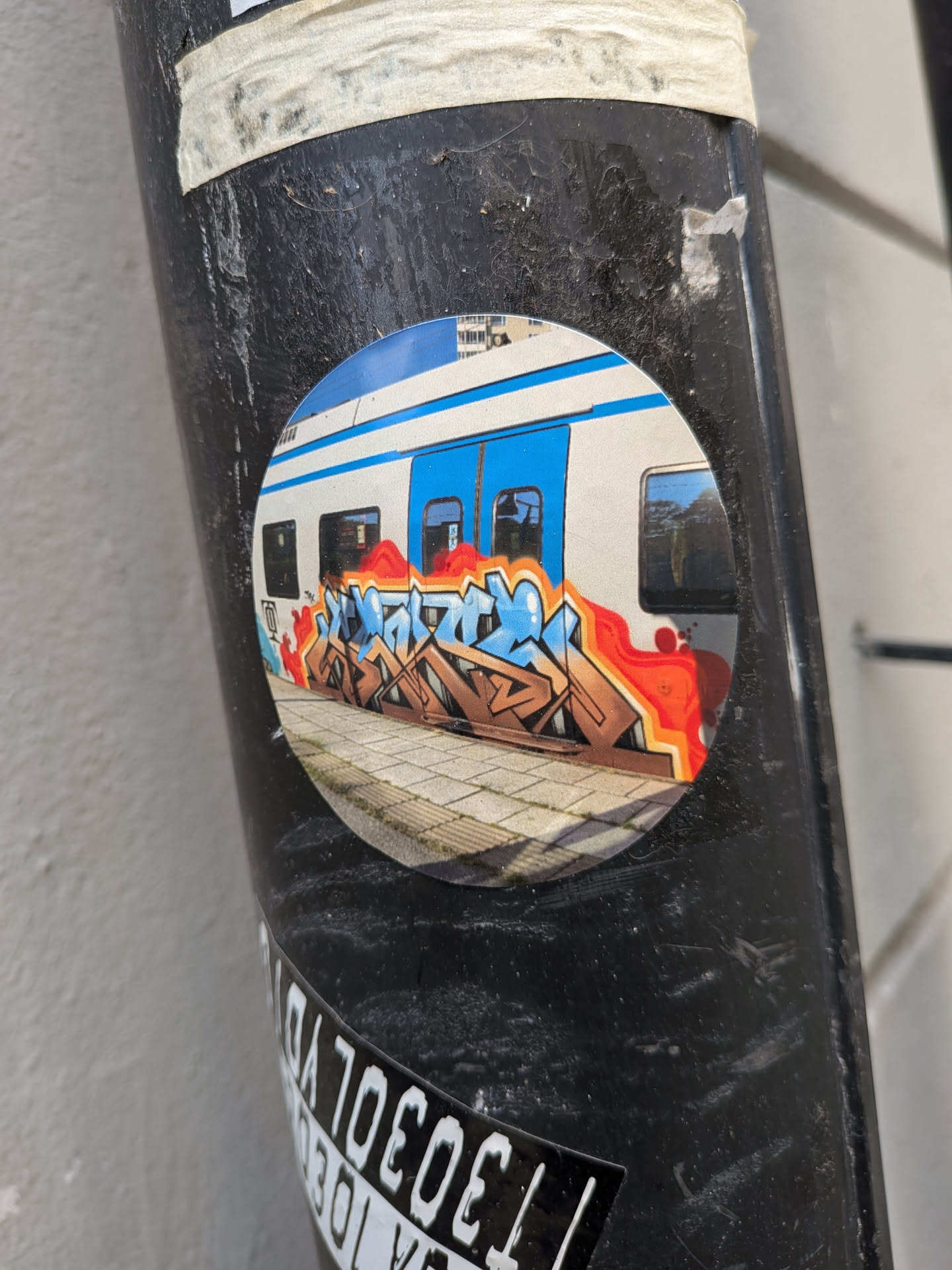 A circular sticker displaying a photograph of a white and blue train car at a platform, decorated with a colorful wildstyle graffiti piece in brown, blue, and orange.