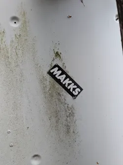 Street sticker A diagonal rectangular sticker with a black background and bold white 'MAKKS' text, stuck on a dirty white surface.