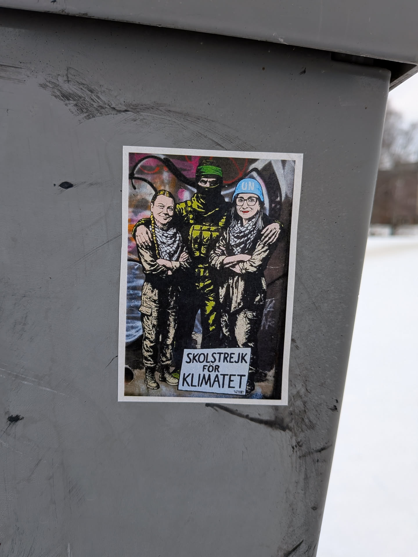 Street sticker A rectangular sticker with a white border, depicting three stylized figures standing side-by-side against a background of abstract graffiti-like shapes in pink, orange, and grey. From left to right, the first figure resembles Greta Thunberg, smiling, wearing a military-style jacket and cargo pants with a patterned scarf. The central figure is taller, masked with a dark balaclava, dressed in camouflage, with arms around the other two. The third figure, resembling Luisa Neubauer, wears glasses, a patterned scarf, a blue beanie with 'UN' written on it, and military-style attire, with arms crossed. At the bottom center, a white sign held by the central figure reads 'SKOLSTREJK FÖR KLIMATET' in black text. The overall aesthetic is gritty and stencil-like, using a limited color palette of greens, browns, black, and white, with accents of blue and background colors.