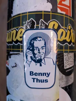 Street sticker The sticker features a stylized blue and white illustration of a man holding a telephone to his ear. He has dark hair, a smiling expression, and is wearing a collared shirt and tie. The background behind his head consists of vertical blue lines. The overall shape of the sticker is a rounded rectangle with a thick blue border. Below the illustration, the name 'Benny Thus' is printed in bold blue capital letters. The sticker is affixed to a surface with a checkered pattern (dark green and yellow) in the background, and other stickers are partially visible around it.