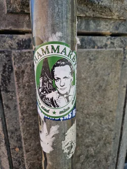A round green and white sticker with the word Hammarby written around the edges. It features a black and white portrait of a man and a drawing of a church.  Some text is partially visible around the portrait, possibly names or a short phrase.