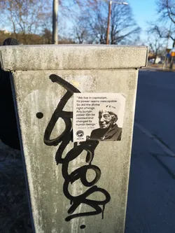 Street sticker A rectangular sticker with a white background, featuring a black and white portrait of Ursula K. Le Guin on the right side. To the left of the portrait, a quote is printed in black text. Below the quote and portrait, there is a small circular logo depicting a stylized figure with a raised fist, and the text 'Allt åt alla' underneath. The sticker is slightly creased and adhered to a weathered, grey metal utility pole.