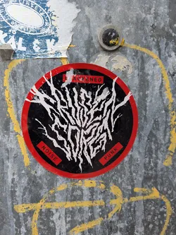 Street sticker A circular sticker with a bold red outer ring enclosing a black inner circle. The central design features an intricate, chaotic pattern of thick white lines, resembling tangled roots, veins, or a lightning strike, spreading across the black background. Three small, rectangular red boxes with white text are positioned around the central design: 'BLACKENED' at the top, 'NOISE' at the bottom left, and 'PUNK' at the bottom right. The sticker shows signs of wear and tear, with slight peeling and scuffs, indicating it's adhered to a textured, possibly metallic, surface.