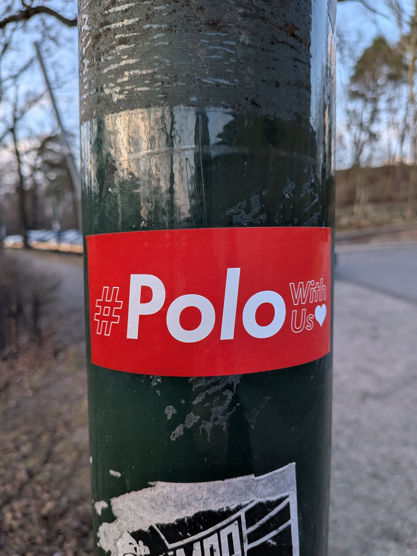 A rectangular sticker with a vibrant red background. The main text, "#Polo", is prominently displayed in large, bold white sans-serif letters. The hashtag symbol is stylized with a double outline. To the right of "Polo", the words "With Us" are stacked vertically in smaller, thinner white sans-serif font, followed by a small white heart emoji. The sticker has a clean, direct, and inviting aesthetic.